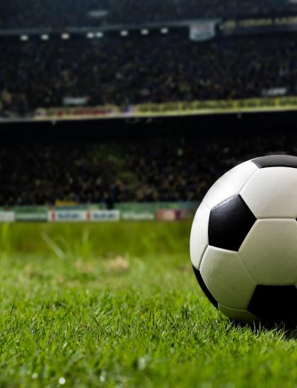 Pallone da calcio su campo verde, stadio affollato sullo sfondo.
