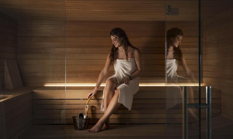 Femme dans un sauna avec seau et louche, enveloppée dans une serviette.